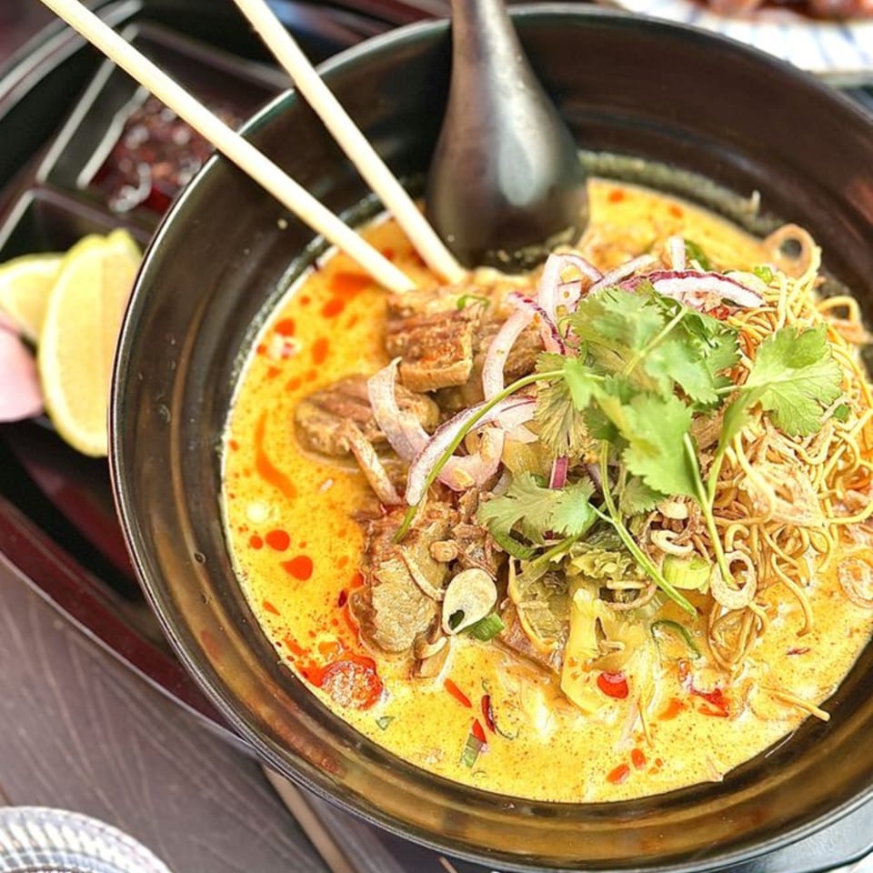 A black bowl of Delicious Thai Kitchen's Khao Soi, a yellow curry noodle soup with meat and herbs.